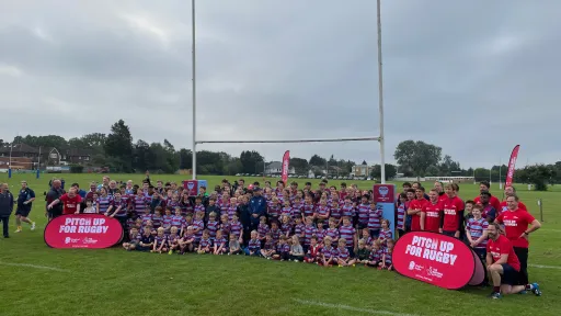 Large group of children and adults posing on a rugby field with goalposts in the background.