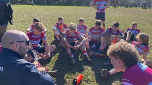 A rugby coach talks to a group of young players sitting on the grass, wearing team jerseys.