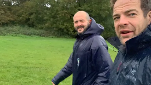 Two volunteers in waterproof jackets walking on a grassy field, with trees in the background.