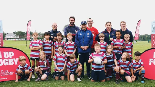 A group of children in rugby shirts poses with coaches on a grassy field, banners in the background.