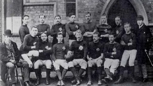 A vintage group photo of a rugby team in dark jerseys, posing in front of a building.