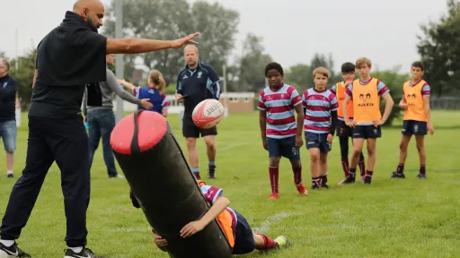A coach demonstrates tackling techniques while young players observe on a rugby field.